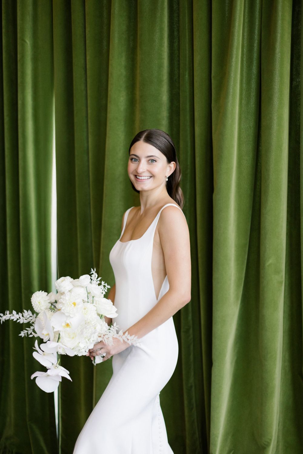 A bride in an elegant white fitted gown holding a white peony bouquet against green velvet curtains.