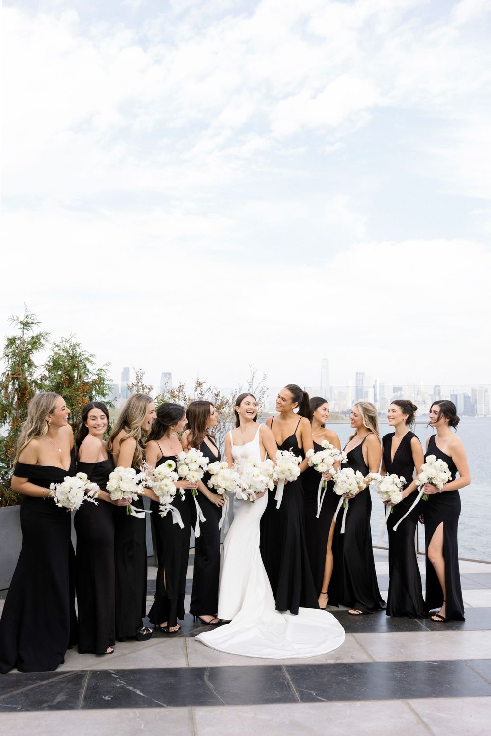 A bridal party in elegant black gowns and a white wedding dress pose together on an outdoor terrace with city views.