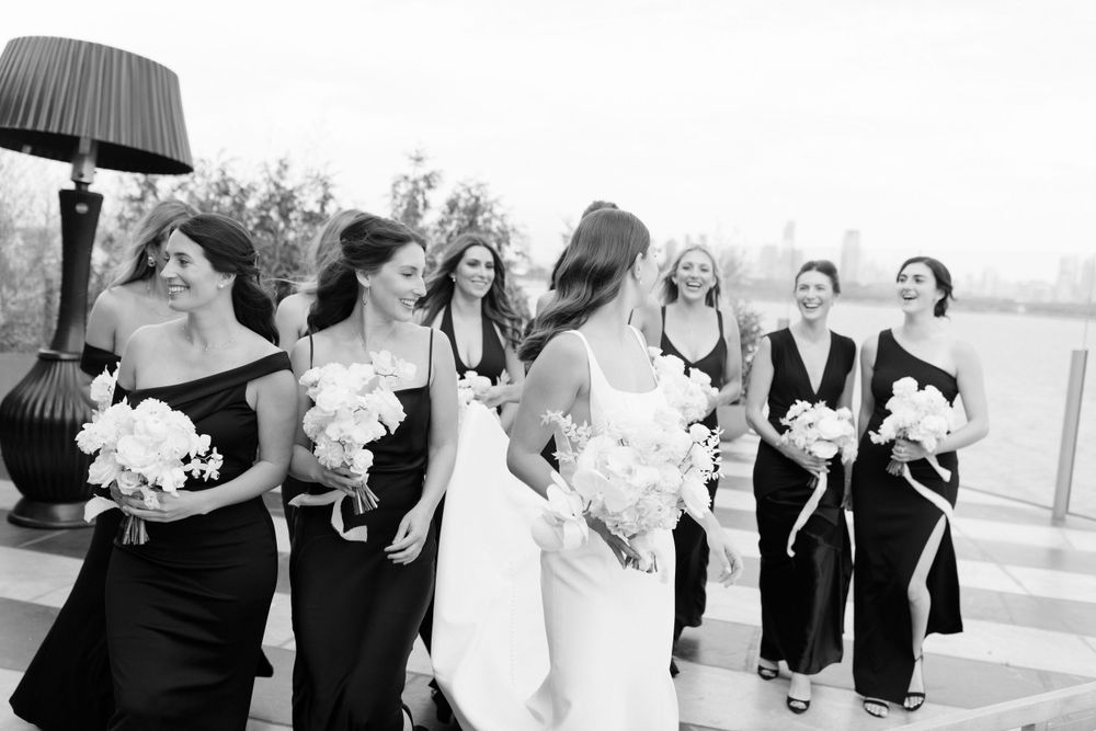 A bridal party in black and white dresses laughs together while holding bouquets outdoors.