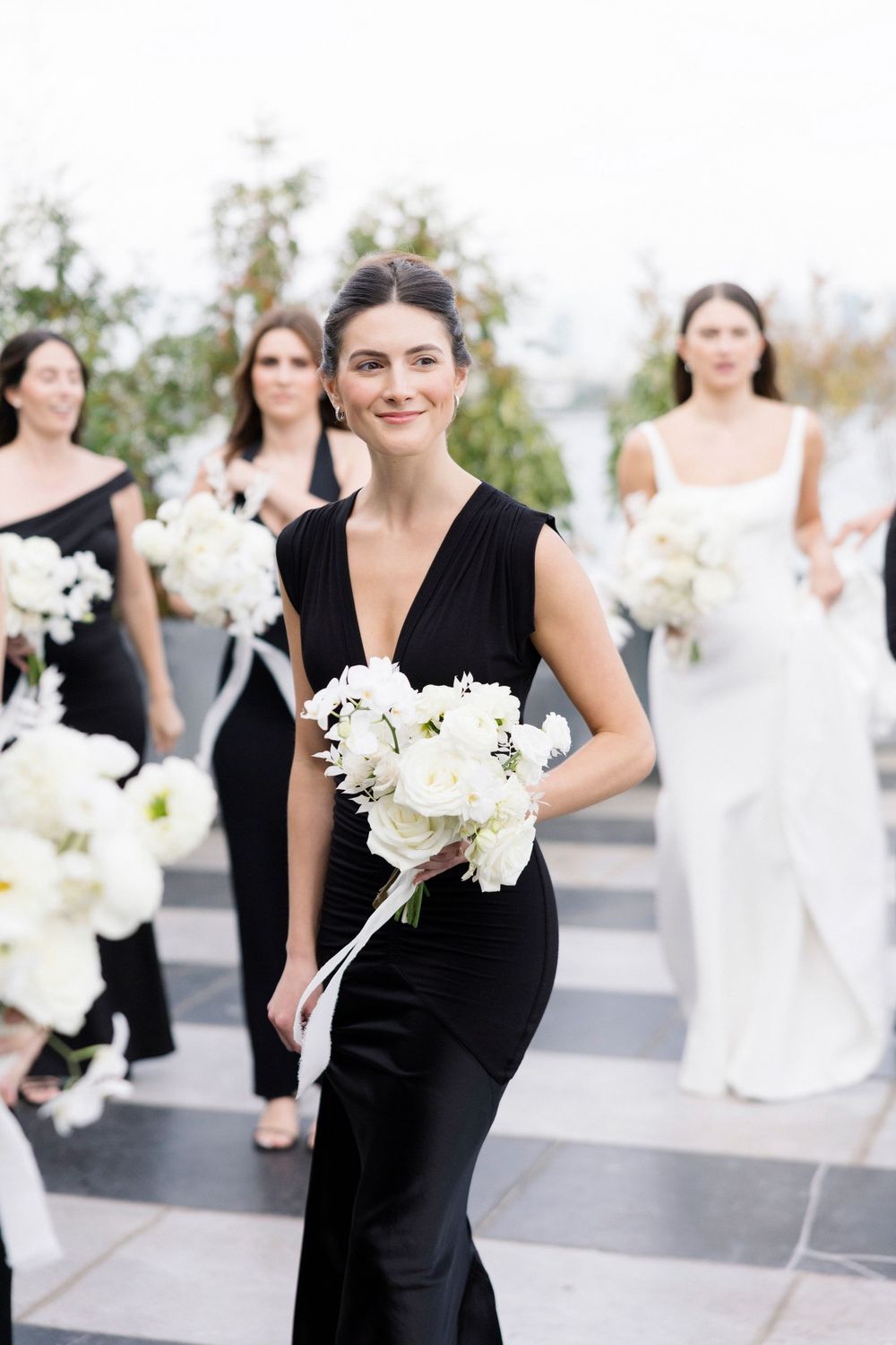 Wedding party members in black and white attire carrying white bouquets on a stone terrace.