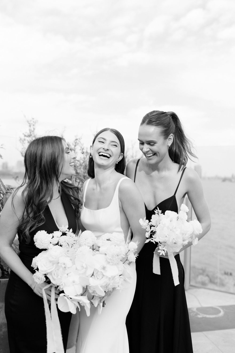 Three women in black and white dresses share joyful laughter while holding elegant white flower bouquets at an outdoor event.
