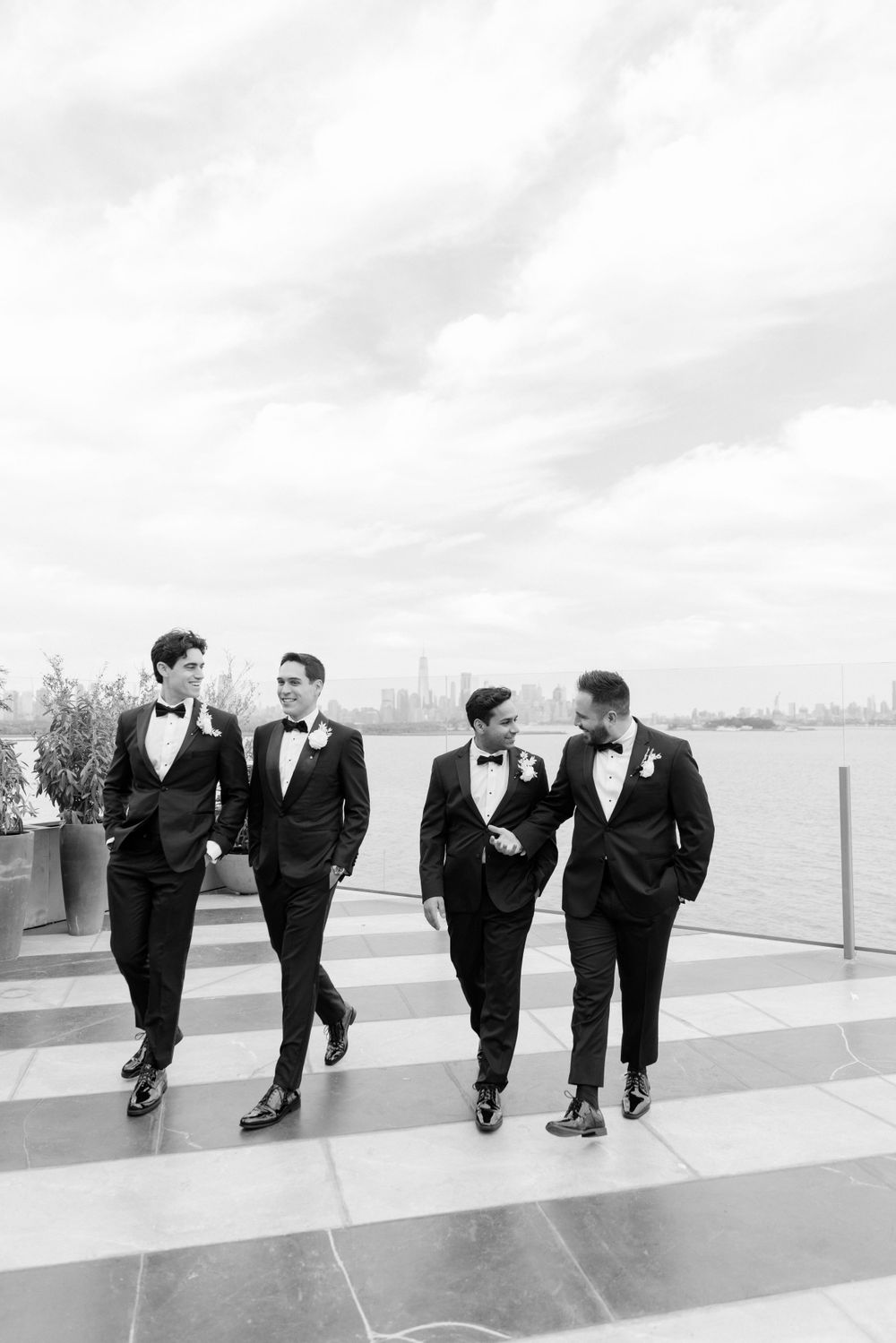 Four groomsmen in classic black tuxedos and bow ties walk along a waterfront terrace in black and white.