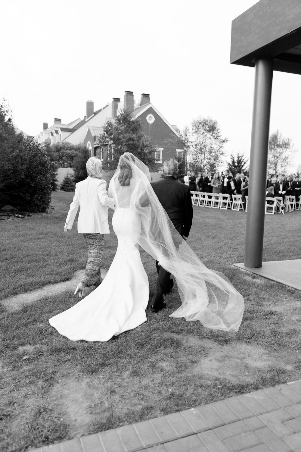 A bride in a flowing white dress and long veil walks outdoors with guests visible in the background at a wedding ceremony.