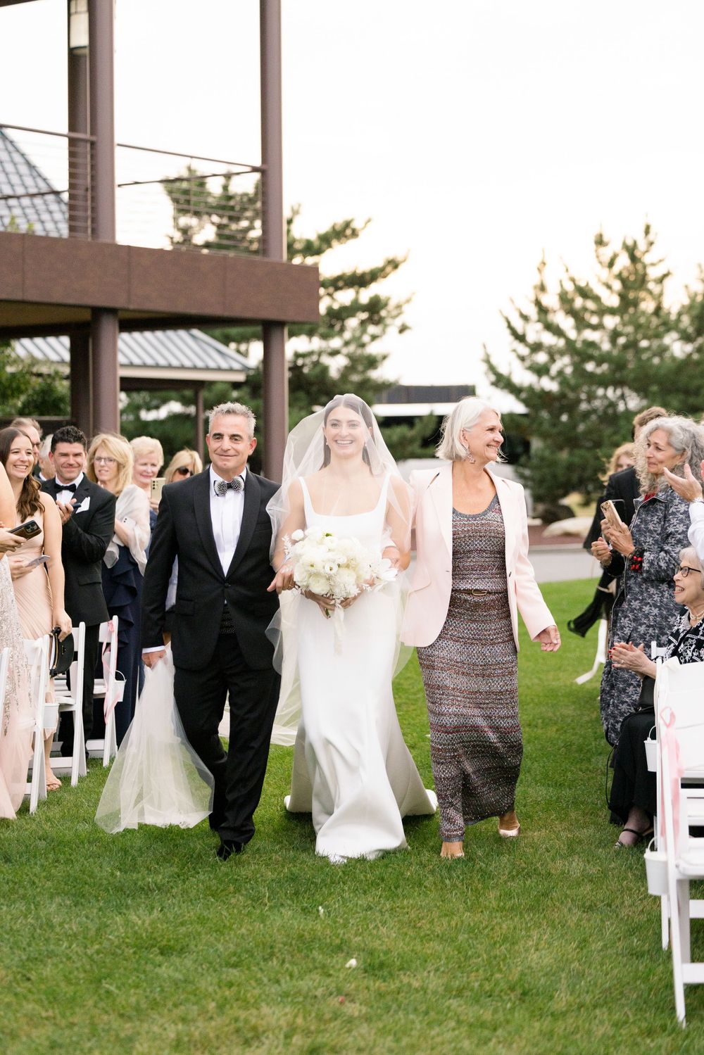 Wedding party walking down the aisle on a green lawn with white chairs during an outdoor ceremony.
