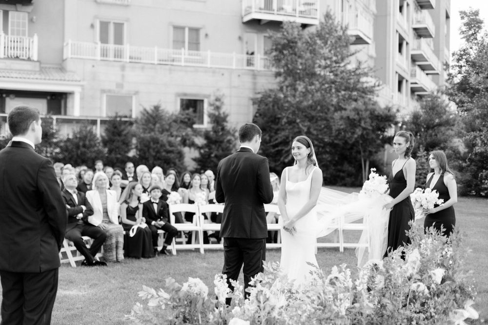 An outdoor wedding ceremony takes place in a garden courtyard surrounded by apartment buildings in black and white.