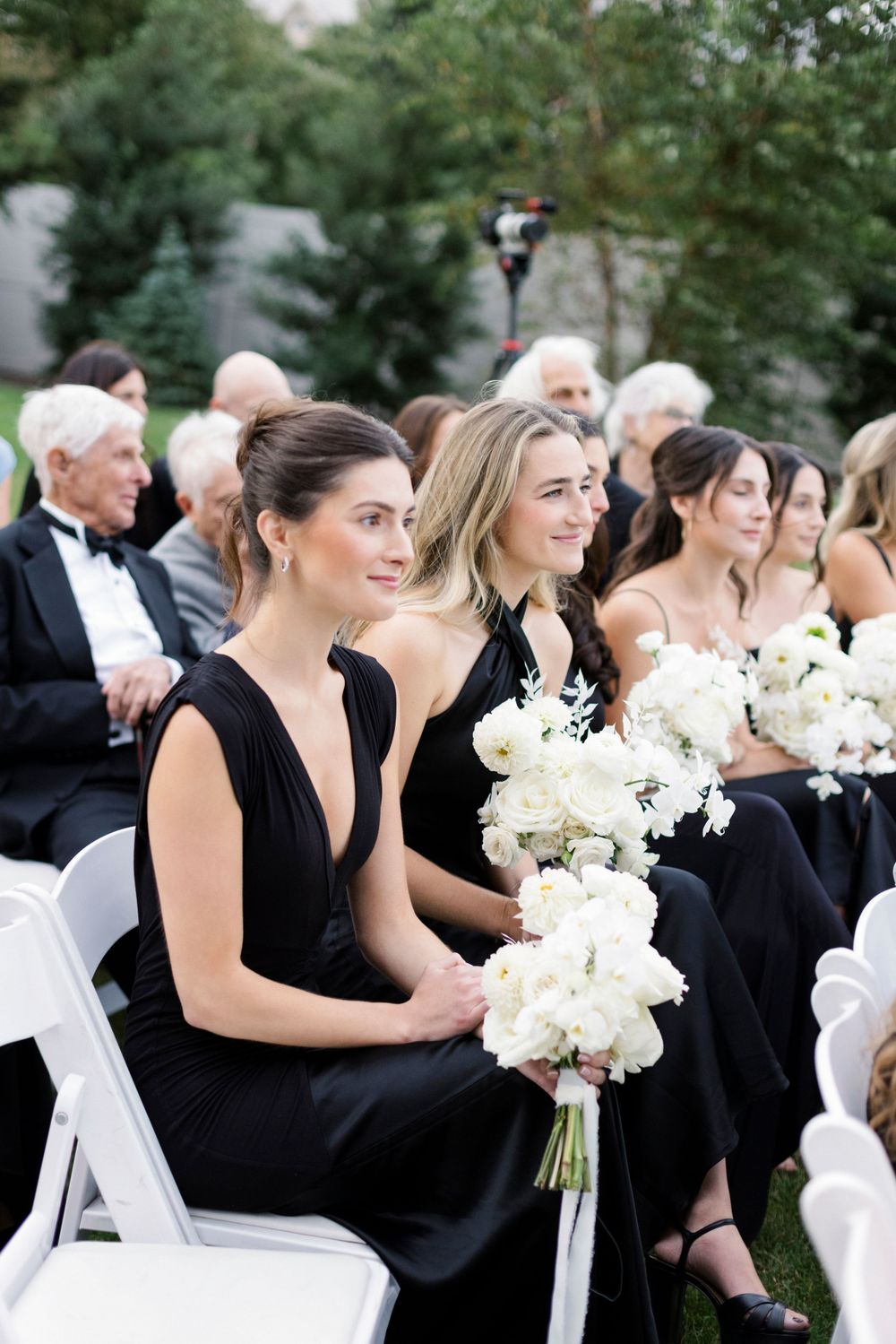 Bridesmaids wearing elegant black dresses hold white bouquets while sitting on white chairs during outdoor ceremony.
