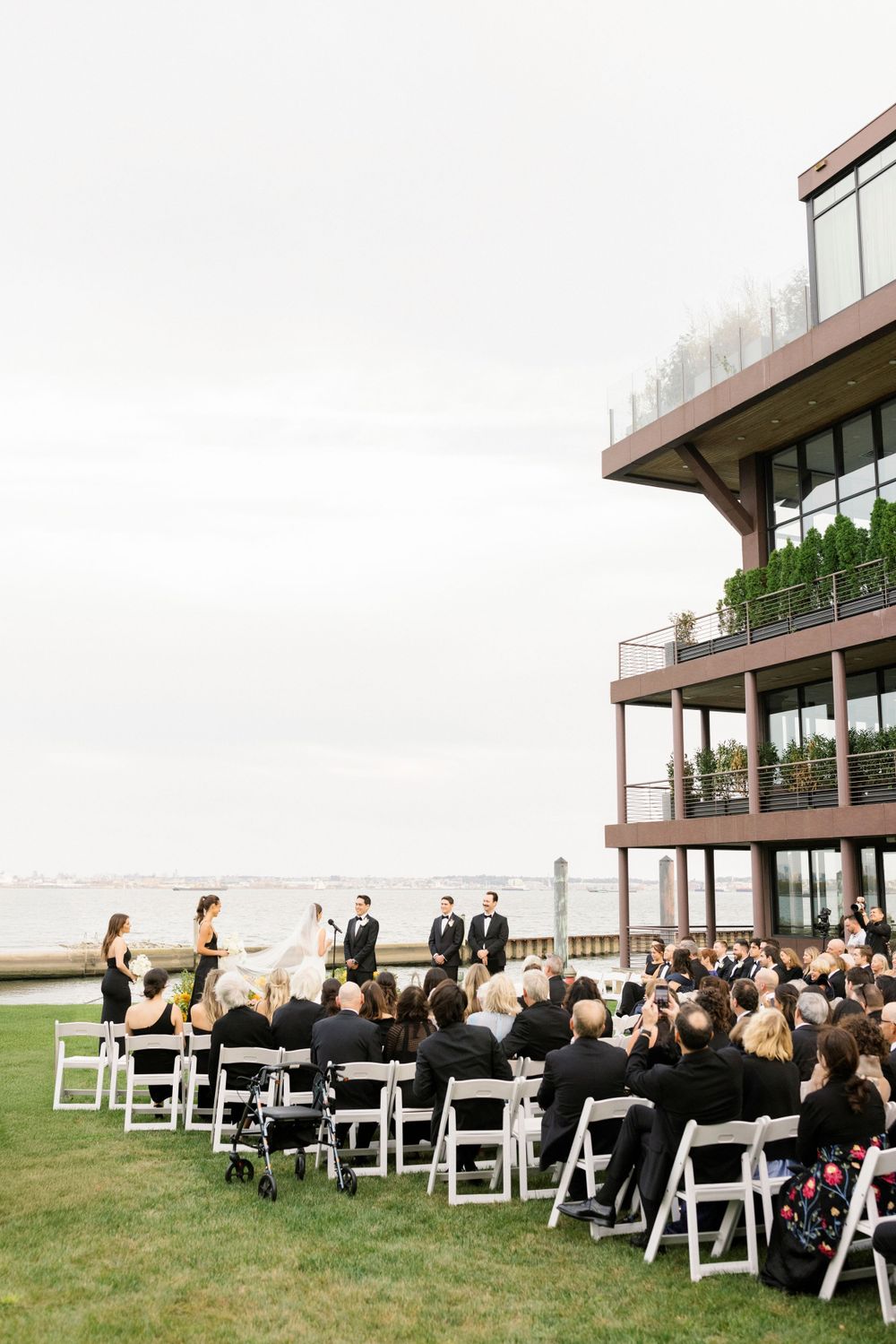 An outdoor wedding ceremony setup with white chairs facing a modern building with waterfront views.