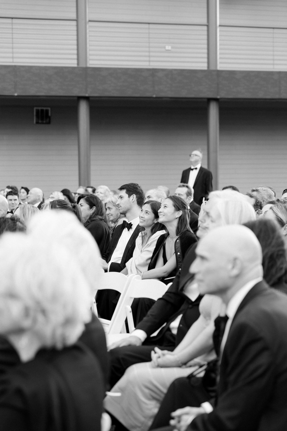 Wedding guests seated in formal attire during outdoor ceremony in black and white.