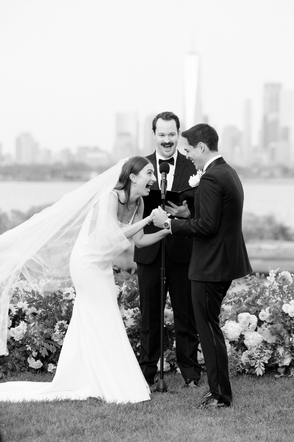 A joyful outdoor wedding ceremony takes place with a city skyline visible in the background as flowers adorn the scene.