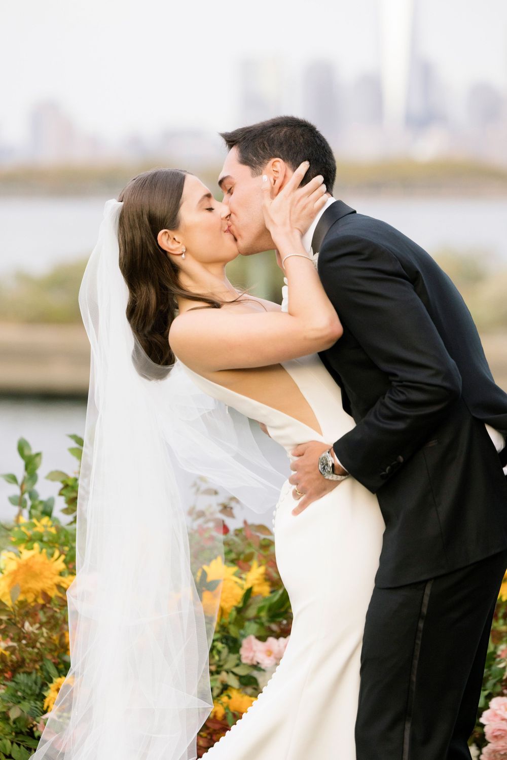 A bride in white gown and groom in black suit share a romantic kiss with yellow flowers and city skyline in background.