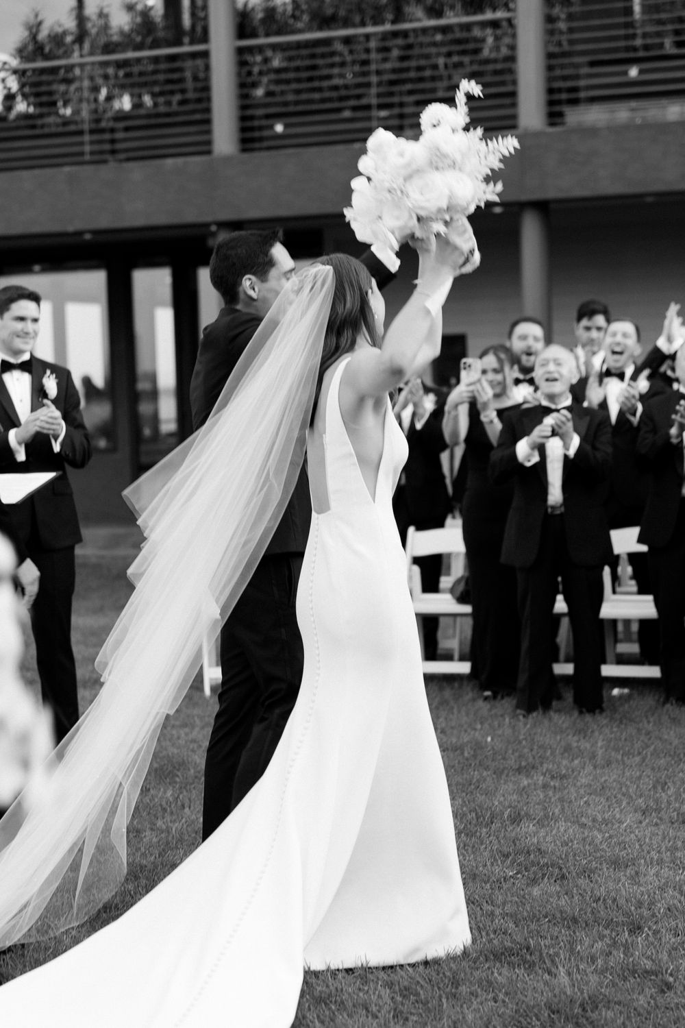 Joyful wedding celebration moment with bride holding bouquet in sleek gown with flowing veil, surrounded by guests.