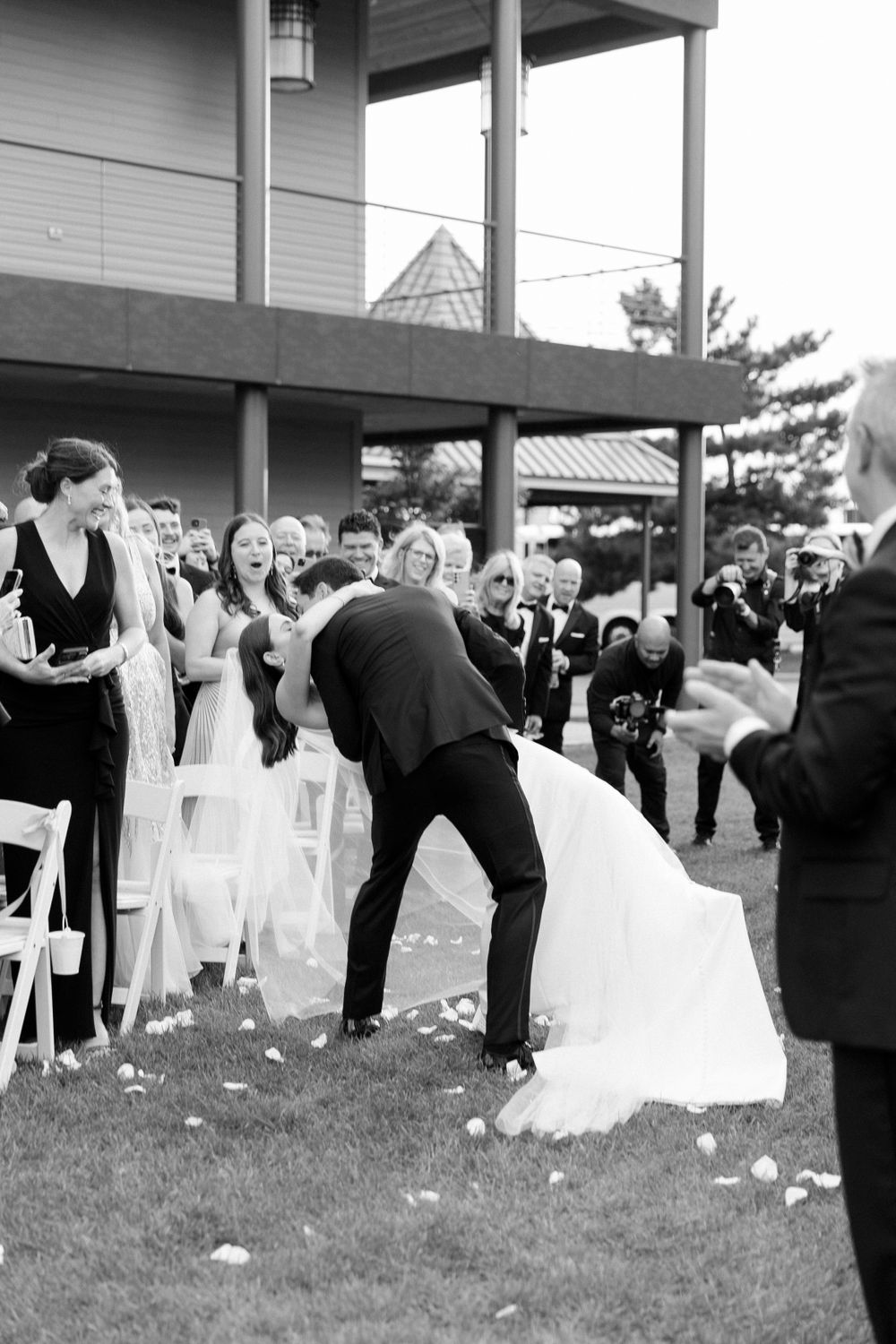 A black and white outdoor wedding ceremony photo showing guests seated on white chairs while rose petals line the ground.