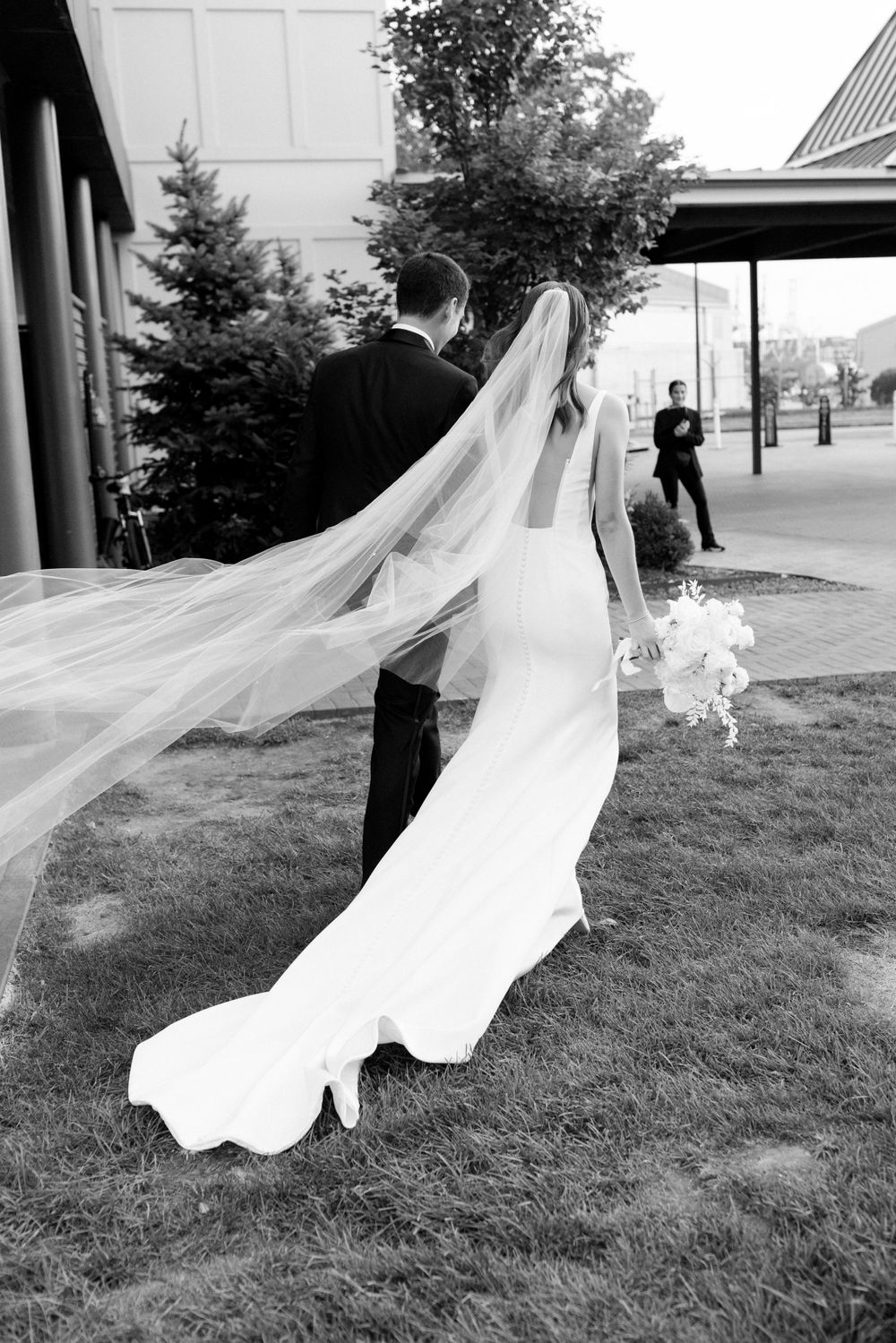 Black and white photo of a wedding couple with flowing veil in an outdoor setting.
