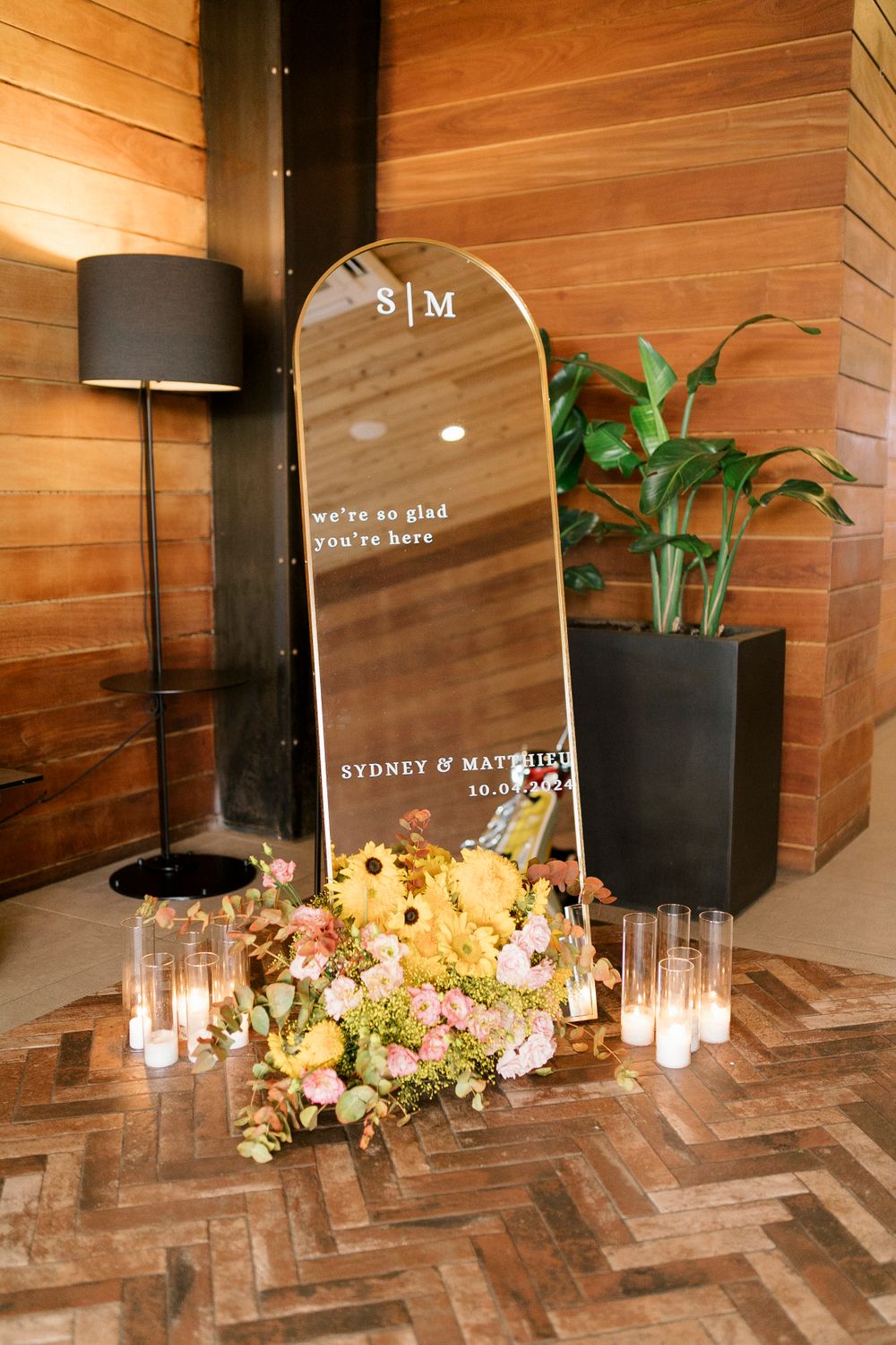 Modern mirror wedding welcome sign with elegant floral arrangements and candles on herringbone wood floor.