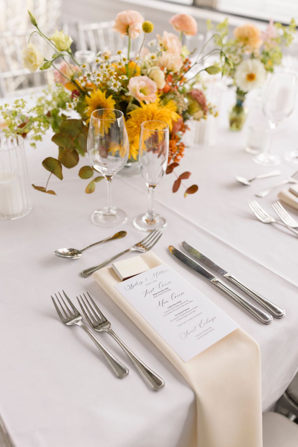 Elegant place setting with menu card, silverware and floral centerpiece at a formal wedding reception table.