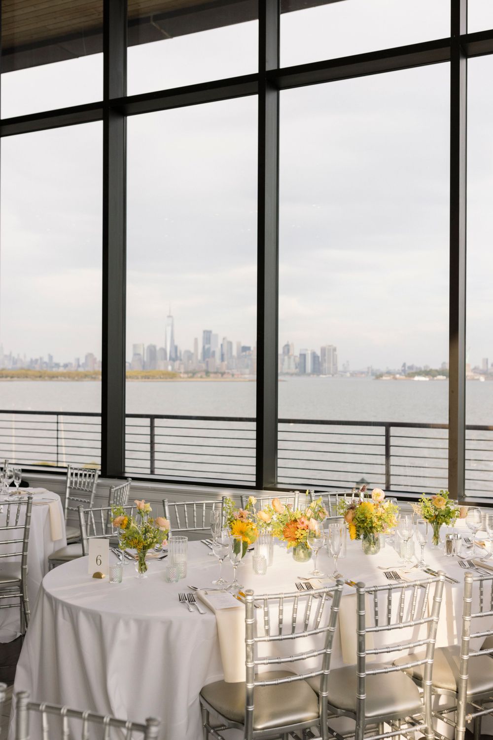 An elegant wedding reception table setting with waterfront city views through floor-to-ceiling windows.