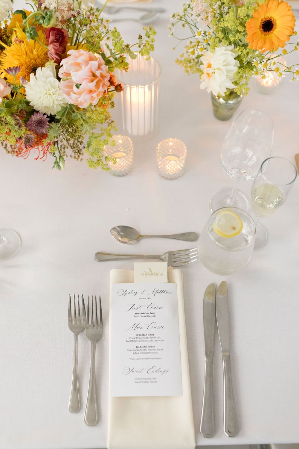 A place setting with menu card, silverware and colorful floral centerpiece on white linen.