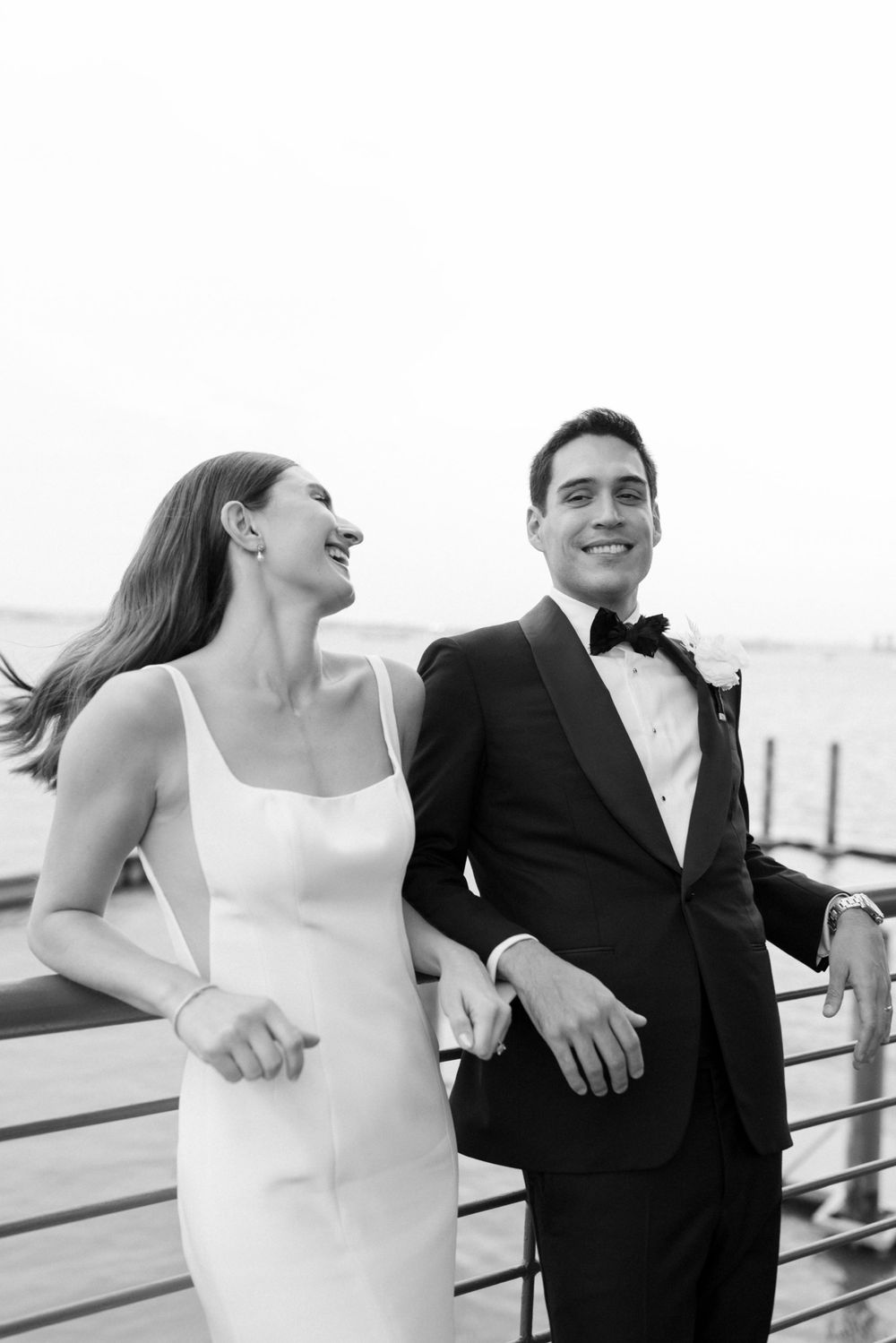 A happy couple smiling together at a waterfront wedding venue, the bride in white and groom in classic tuxedo.