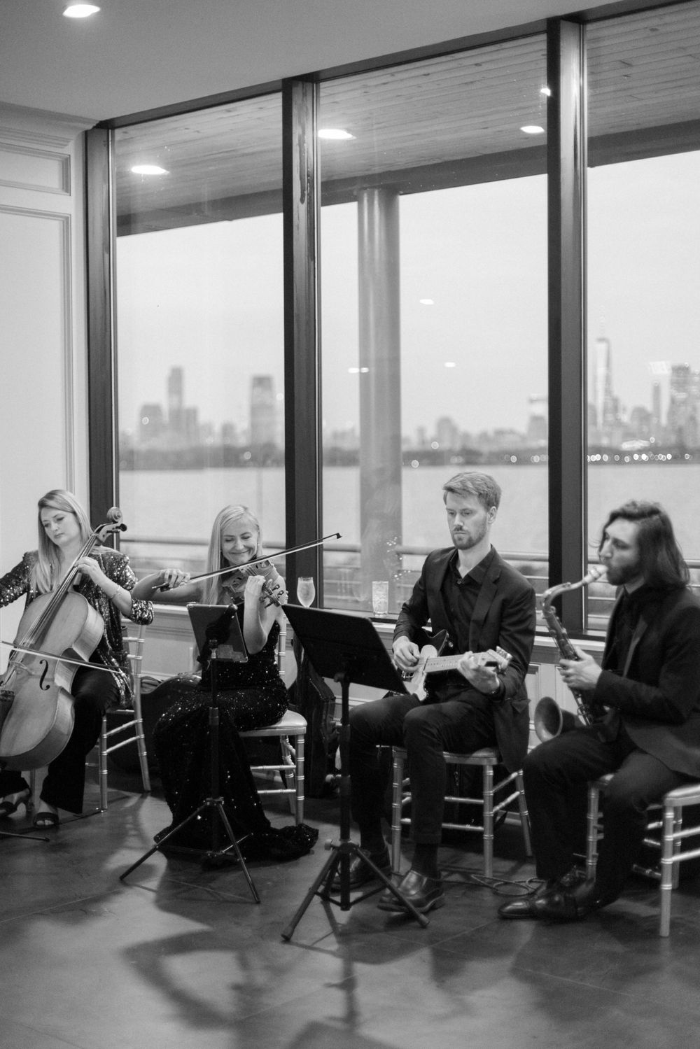A musical quartet performs near large windows overlooking a city skyline in black and white.