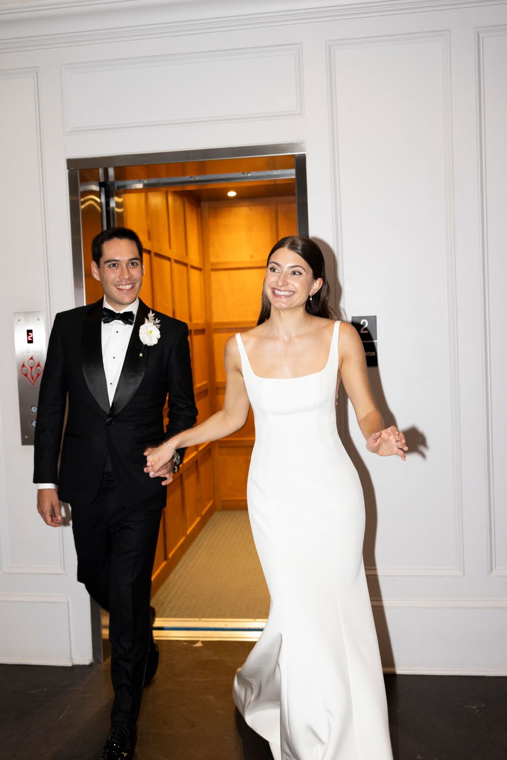 A newlywed couple emerges from an elevator into an elegant hallway with white paneled walls.