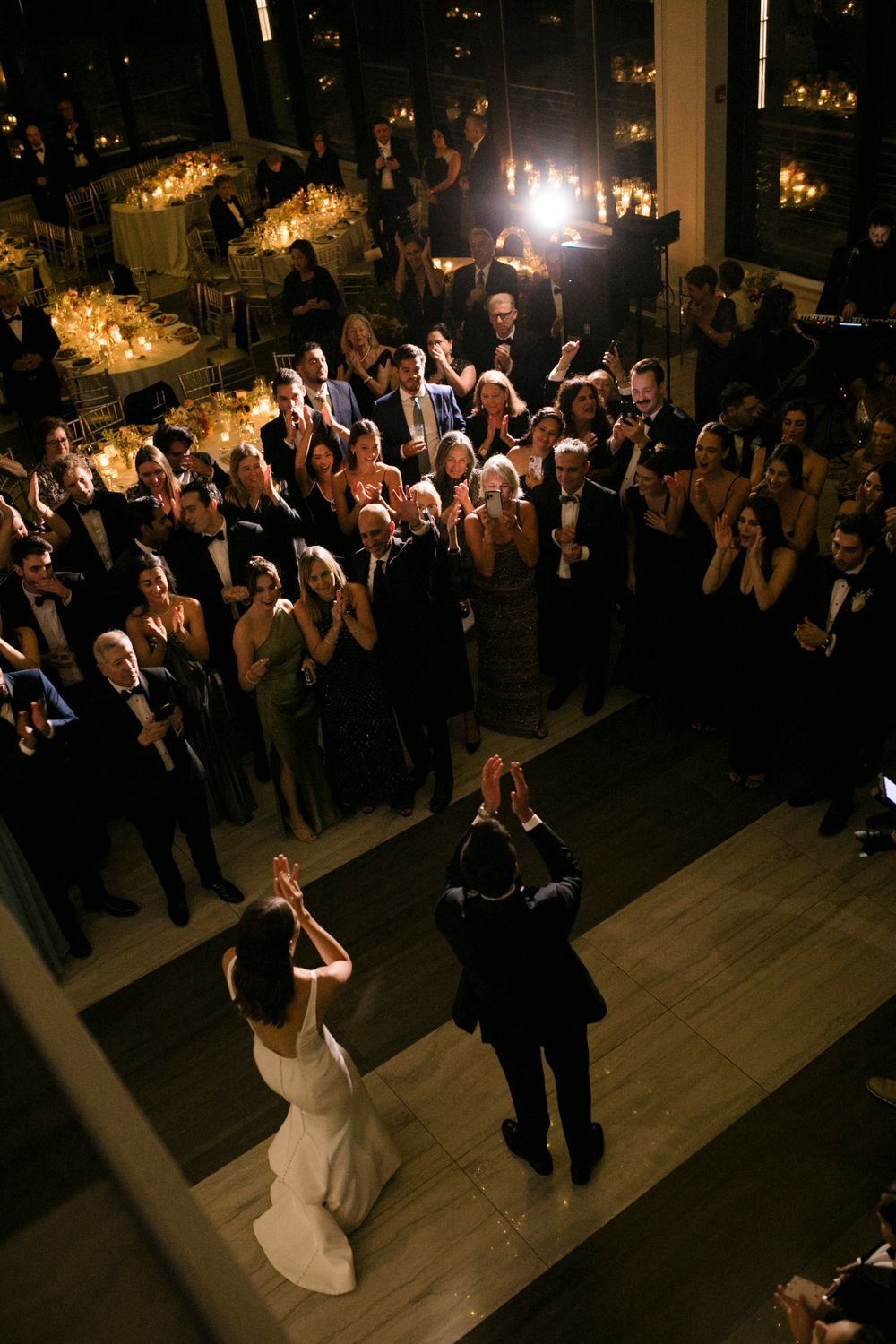 A wedding couple dances on a wooden floor surrounded by elegantly dressed guests in a dimly lit reception venue.