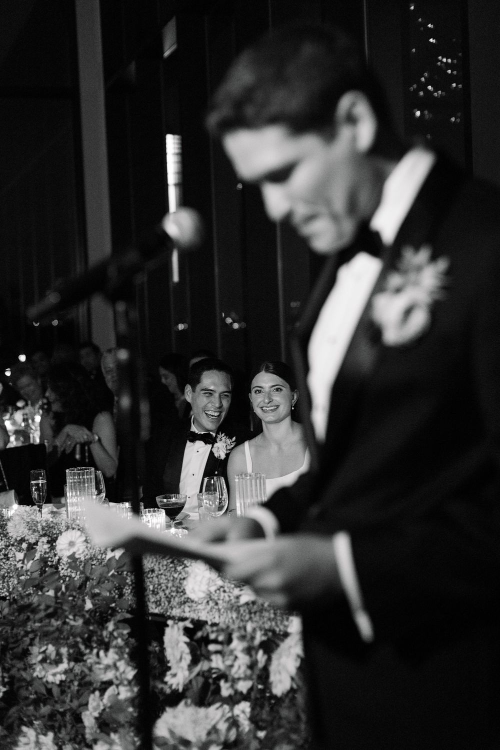 A black and white photo of guests watching a speech at an elegant wedding reception with floral arrangements.