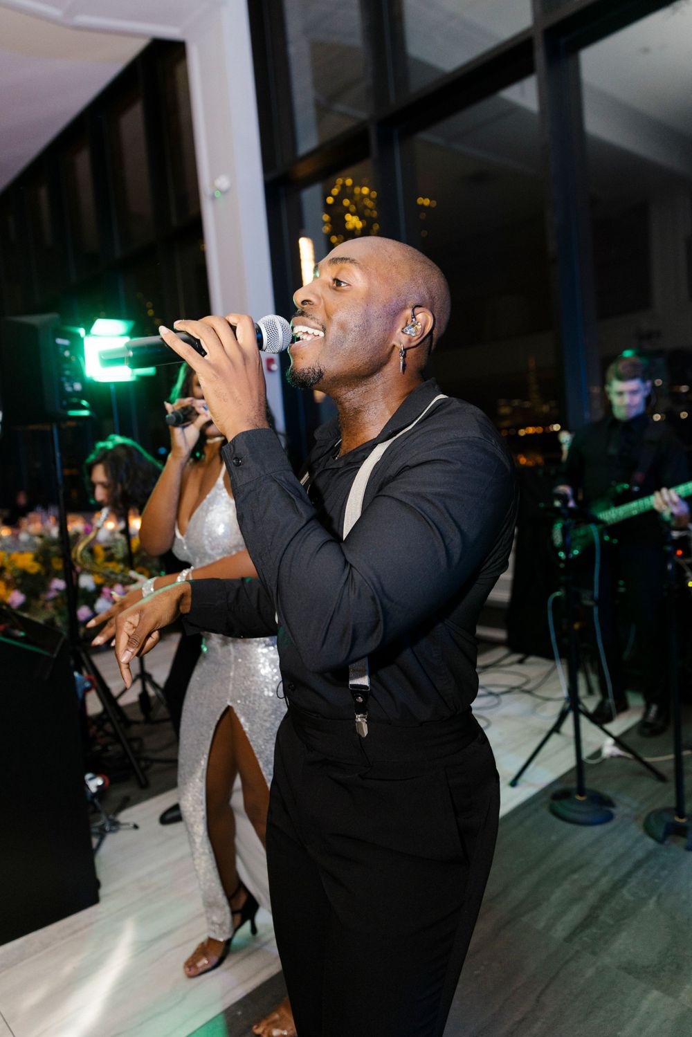 A performer in a black suit sings passionately into a microphone at an indoor event with stage lighting in the background.