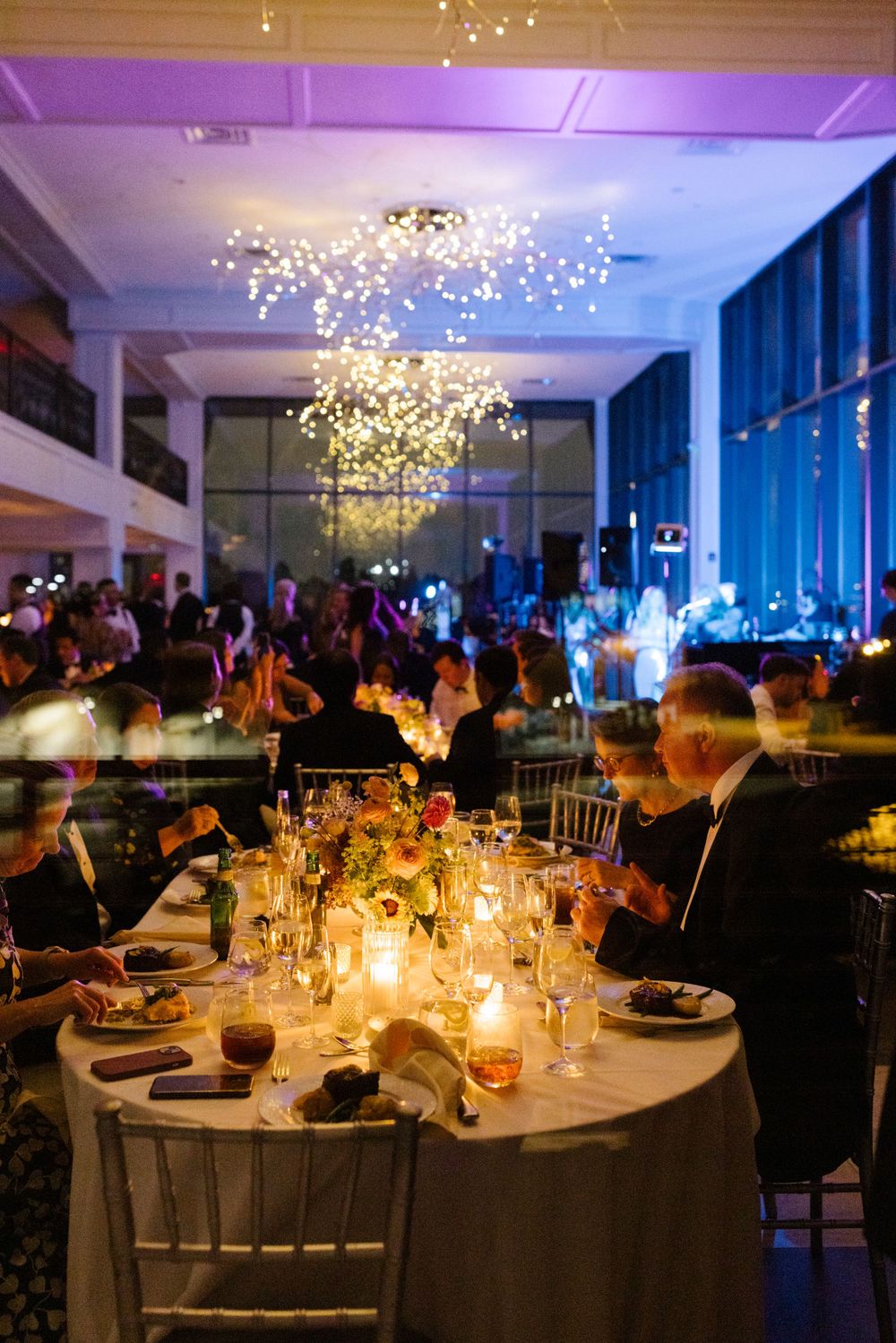 Elegant wedding reception table with candlelight and a dramatic crystal chandelier in a modern venue with blue lighting.