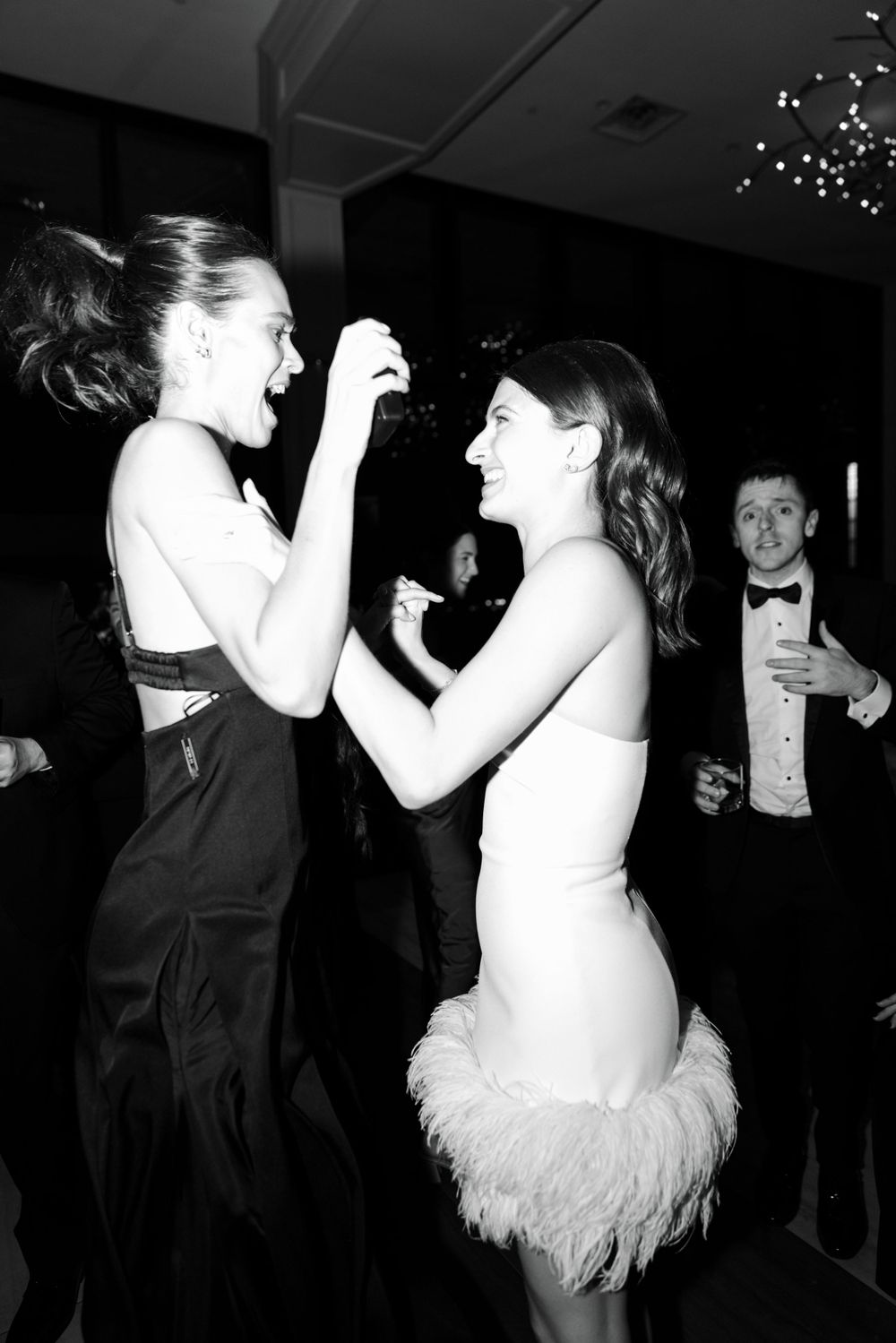 Two women dancing and laughing together at a formal event, one wearing a feathered dress as someone watches in background.
