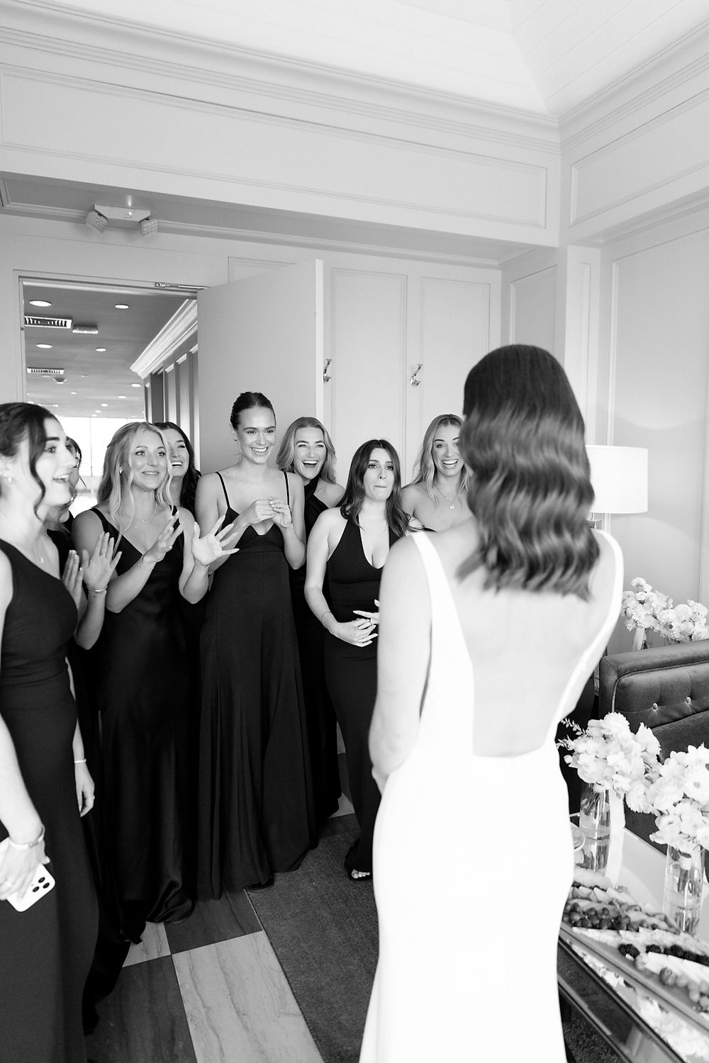 A bridal party in black dresses reacts with excitement during a wedding day first look moment.