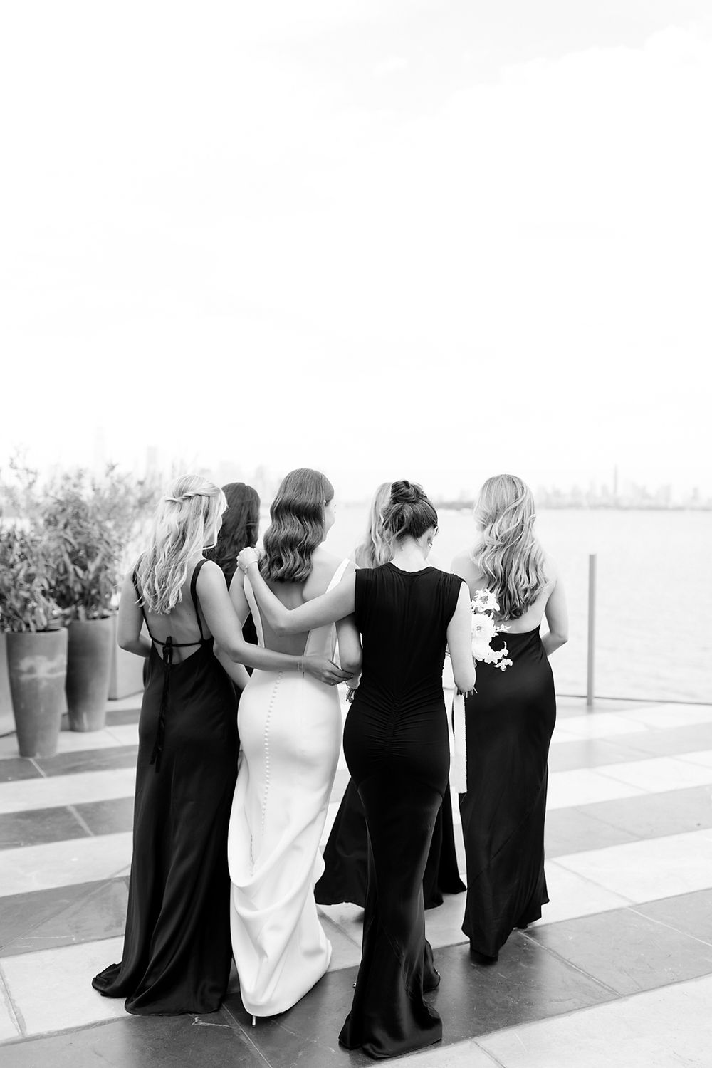 Black and white photo of bridesmaids in long black dresses embracing on a terrace with water views.