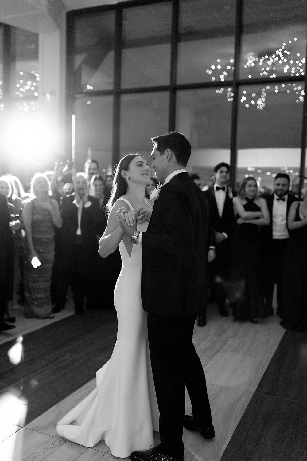 Elegant black and white wedding first dance moment captured with guests watching in a venue illuminated by string lights.
