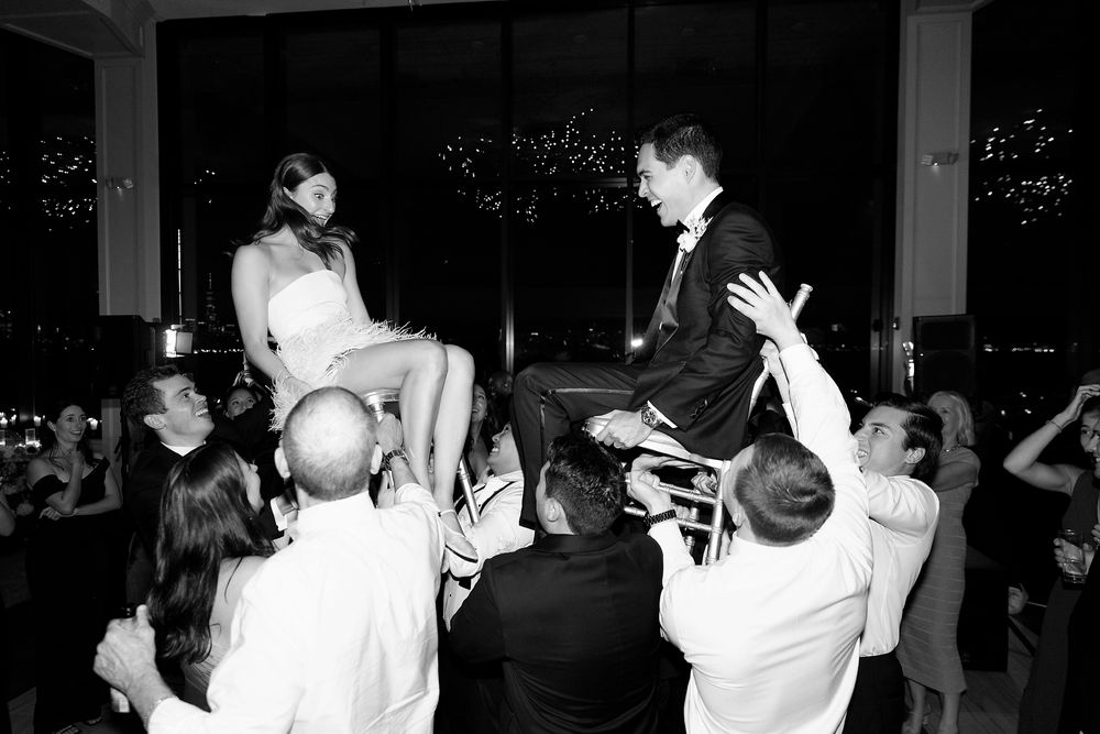 Wedding celebration dancers lift bride and groom in chairs during traditional Hora dance at reception with city lights in background.