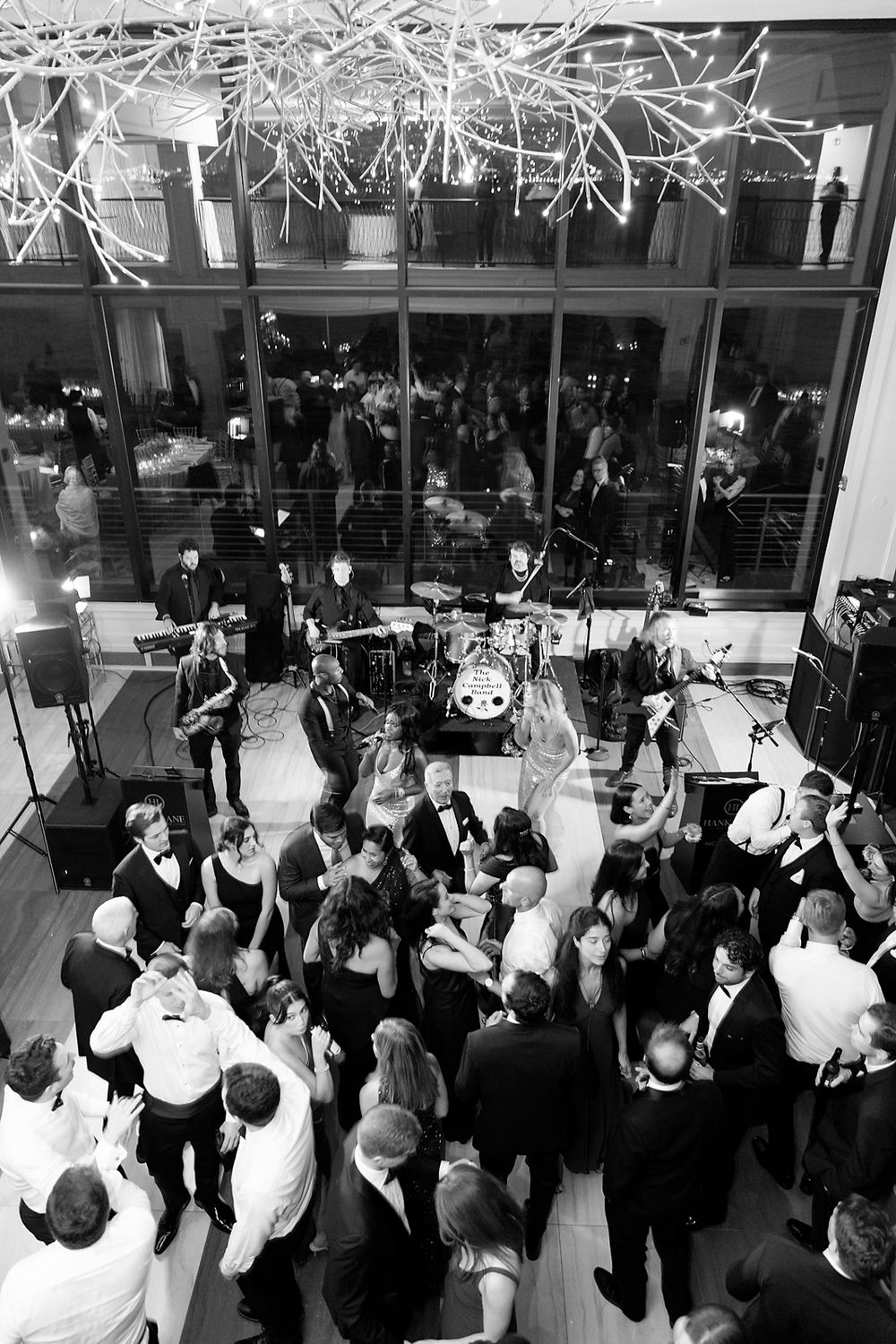 Black and white photo of a lively wedding reception with dancing guests under modern ceiling decor.