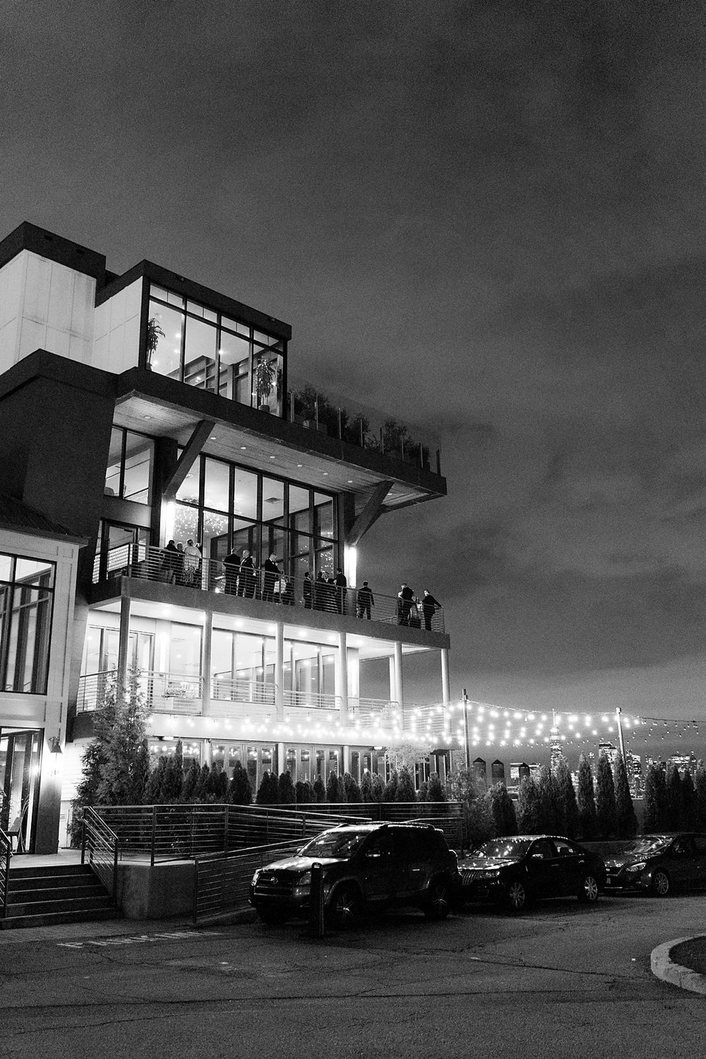 Modern multi-level building lit up at night with string lights and cars parked outside in black and white photography.