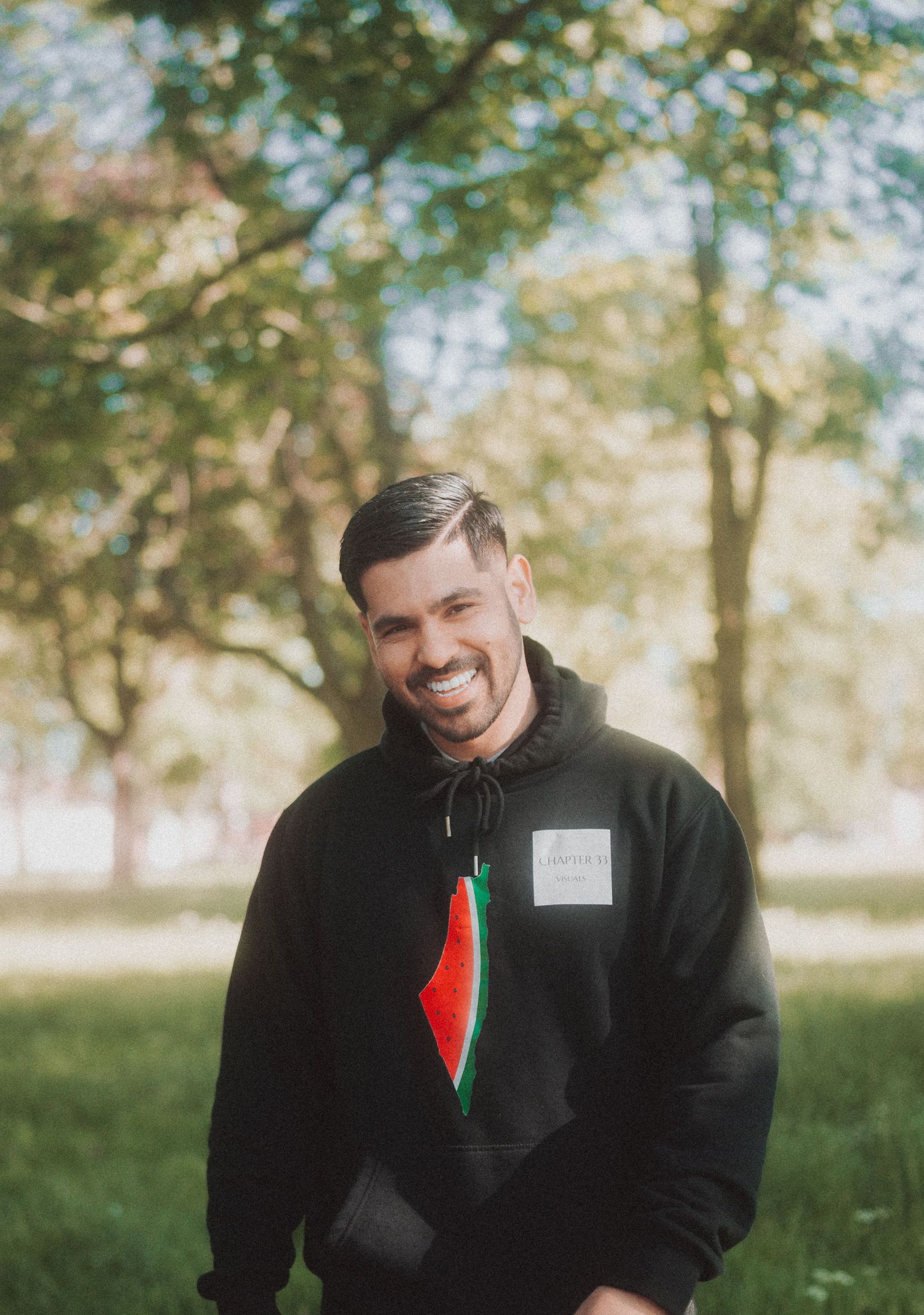 Person wearing a black hoodie with a decorative pin smiling outdoors in a park setting.