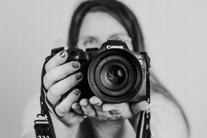 A black and white photo of someone holding a Canon DSLR camera with a large lens pointed directly at viewer.