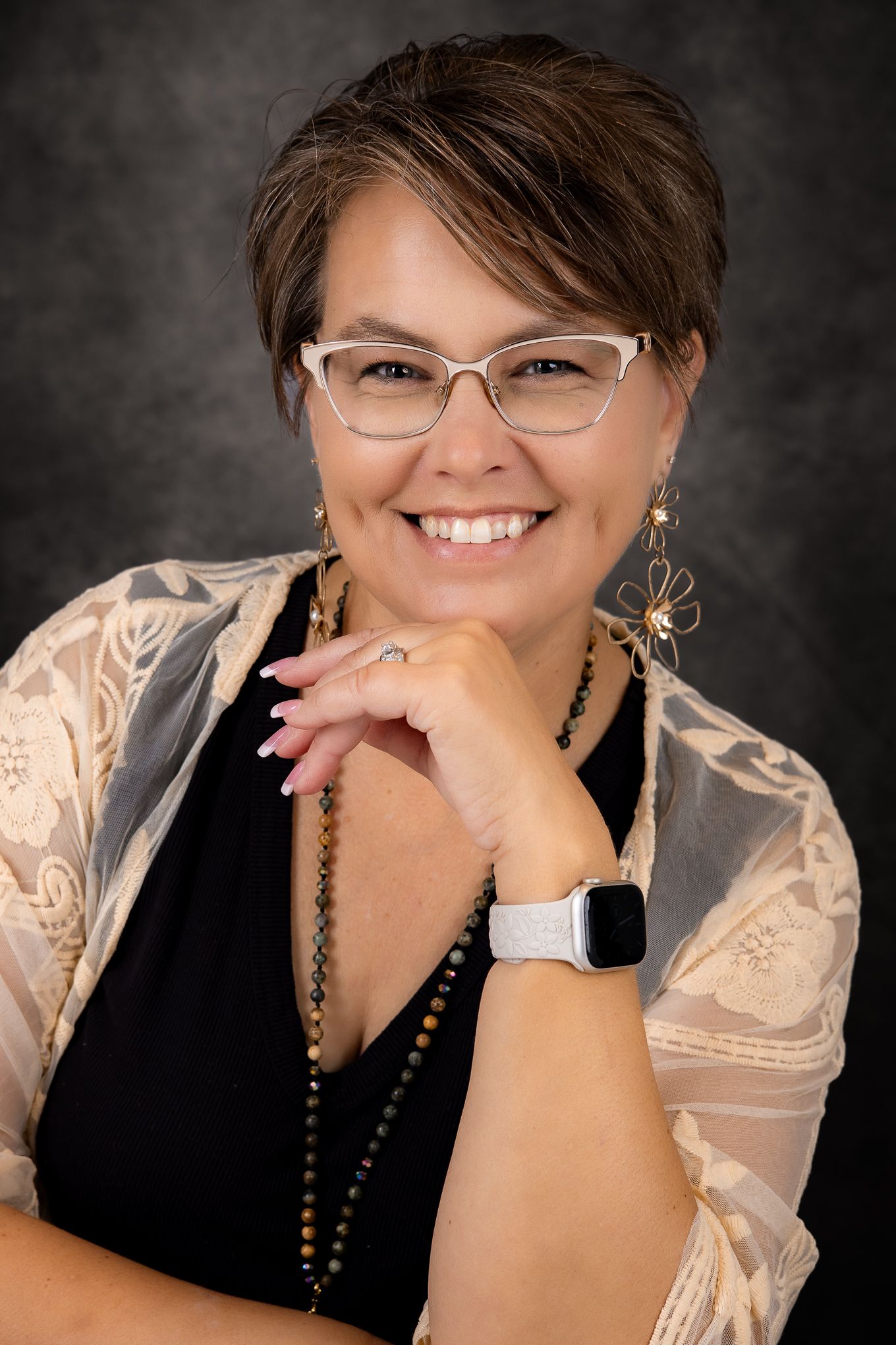 Professional studio portrait with cat-eye glasses, long beaded necklace, and smart watch against dark background.