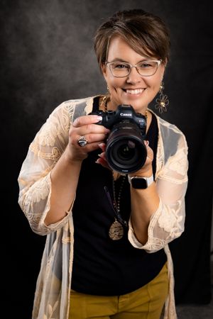 Professional photographer wearing glasses and holding a mirrorless camera smiles while wearing yellow pants and lace cardigan.