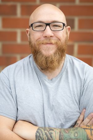 Portrait of a bald man with a full ginger beard wearing glasses and a grey t-shirt against a brick wall background.