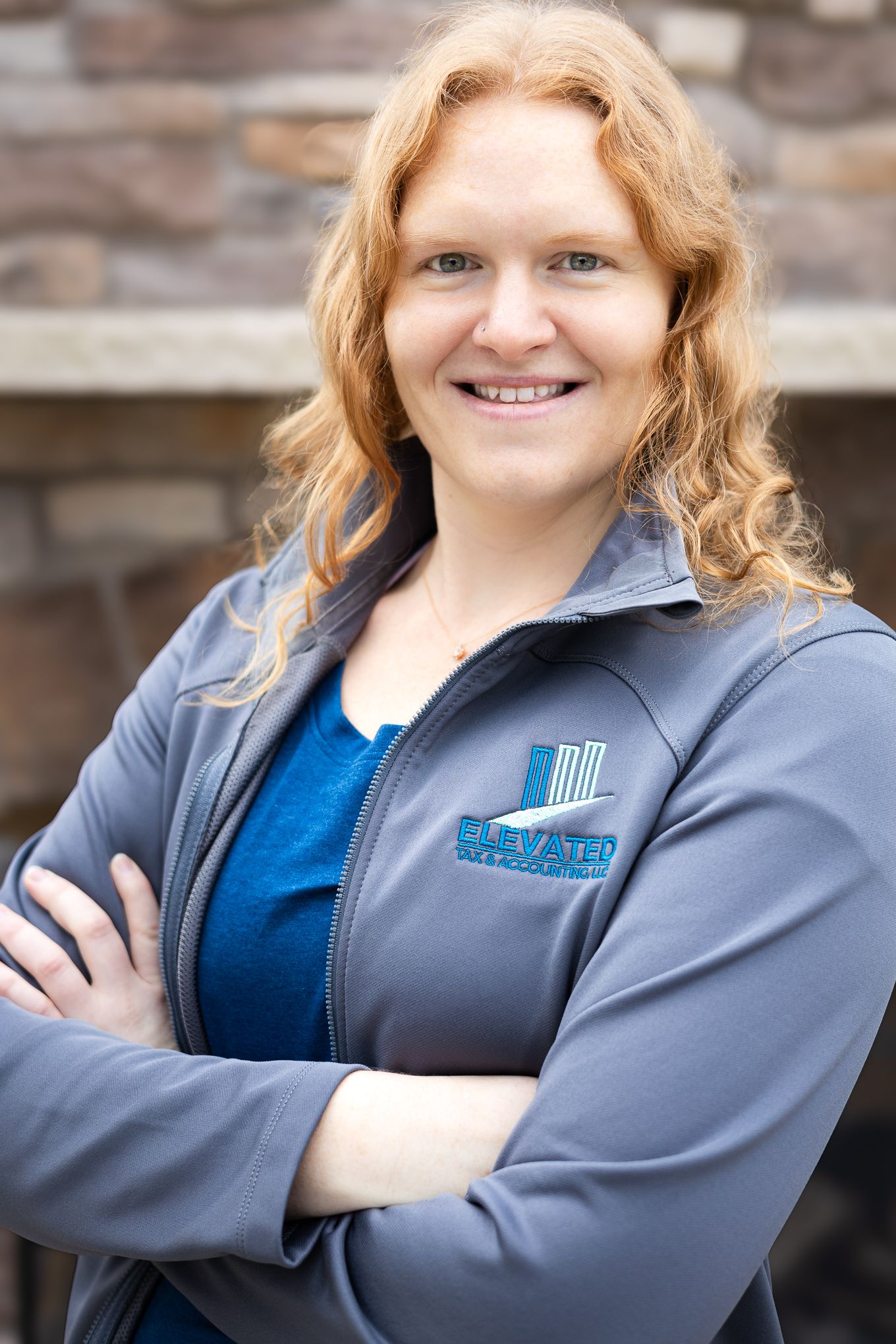 Professional headshot of person in gray jacket with blue shirt and company logo against stone wall background.