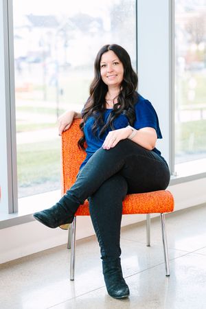 Person in blue top and dark jeans sits casually on an orange chair by bright windows in modern interior space.