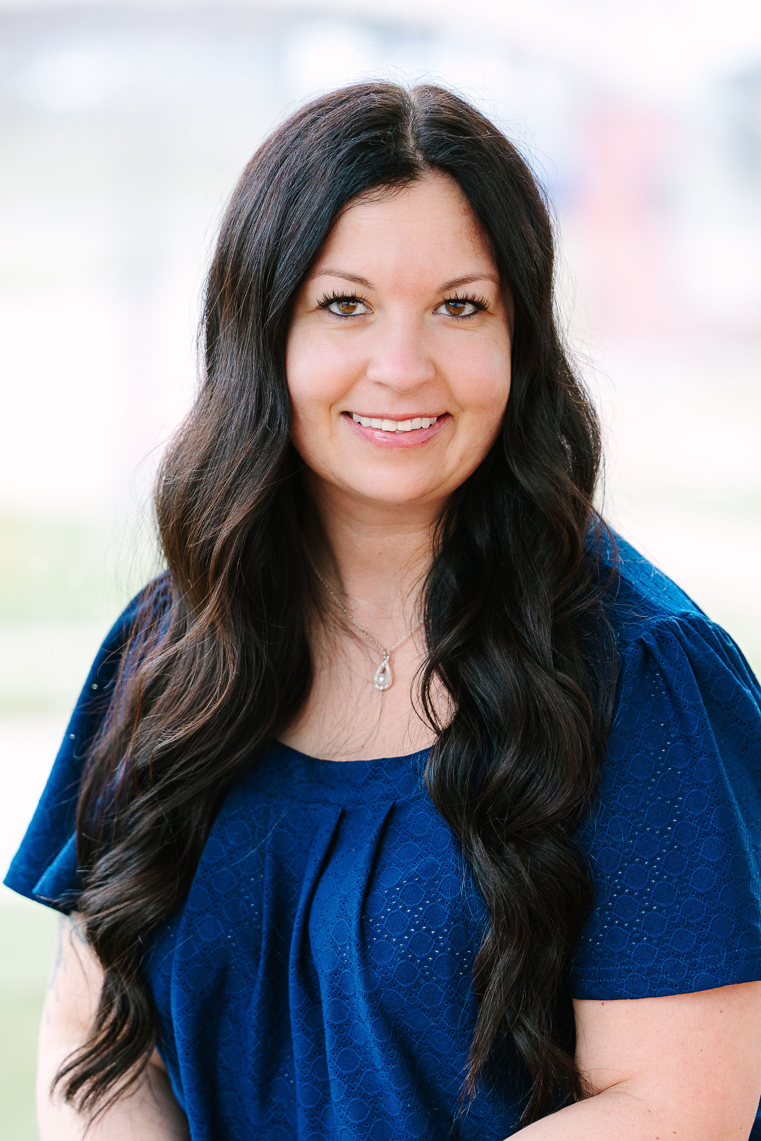 Professional headshot of a person with long dark wavy hair wearing a blue top against a light background.