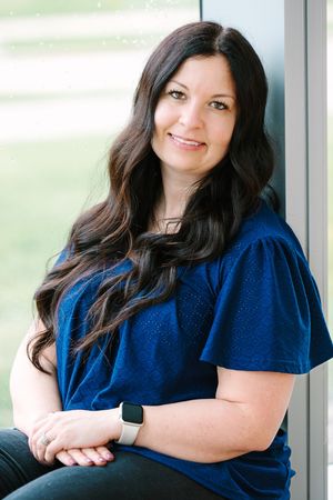 Portrait photo showing a woman with long dark hair wearing a navy blue flutter sleeve top, seated by a window.