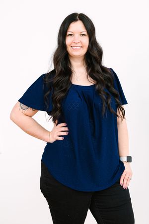 Woman wearing navy blue blouse and black pants poses in casual business attire against white background.