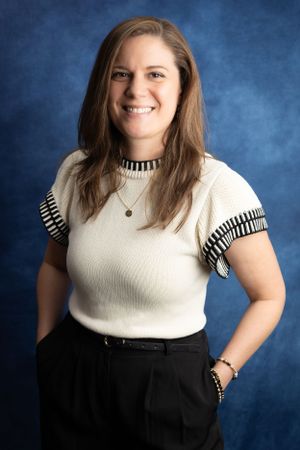 Professional portrait against blue background showing person in cream knit top with striped trim and dark pants.