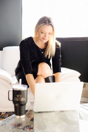 Woman in black sweater working on laptop from home with travel mug on coffee table.