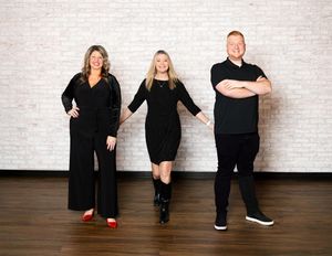 Three people in all-black outfits pose confidently against a white brick wall in a studio setting.