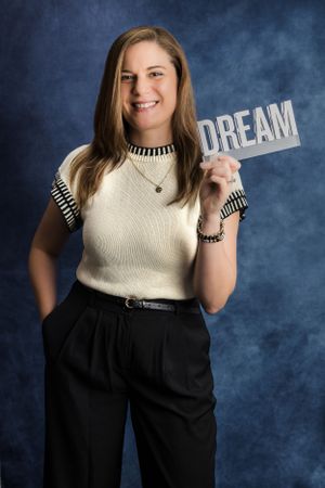 Portrait of a person in black pants and white top holding up a DREAM sign against a blue background.