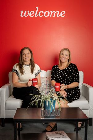 Two people sit on a white couch holding red coffee mugs with a welcome sign and plant on the coffee table.